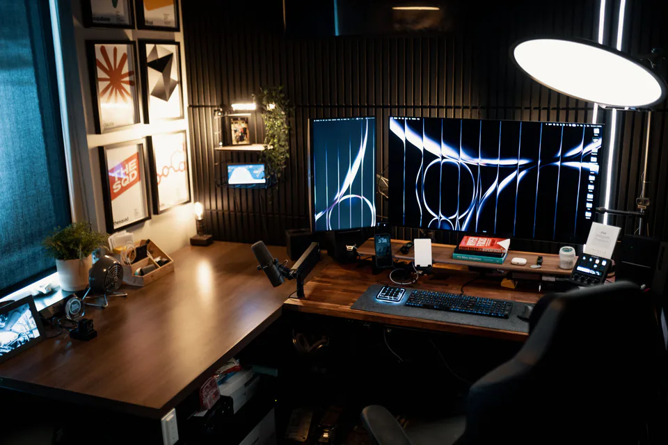 A dark, moody desk setup with a large monitor, microphone and desk lamp