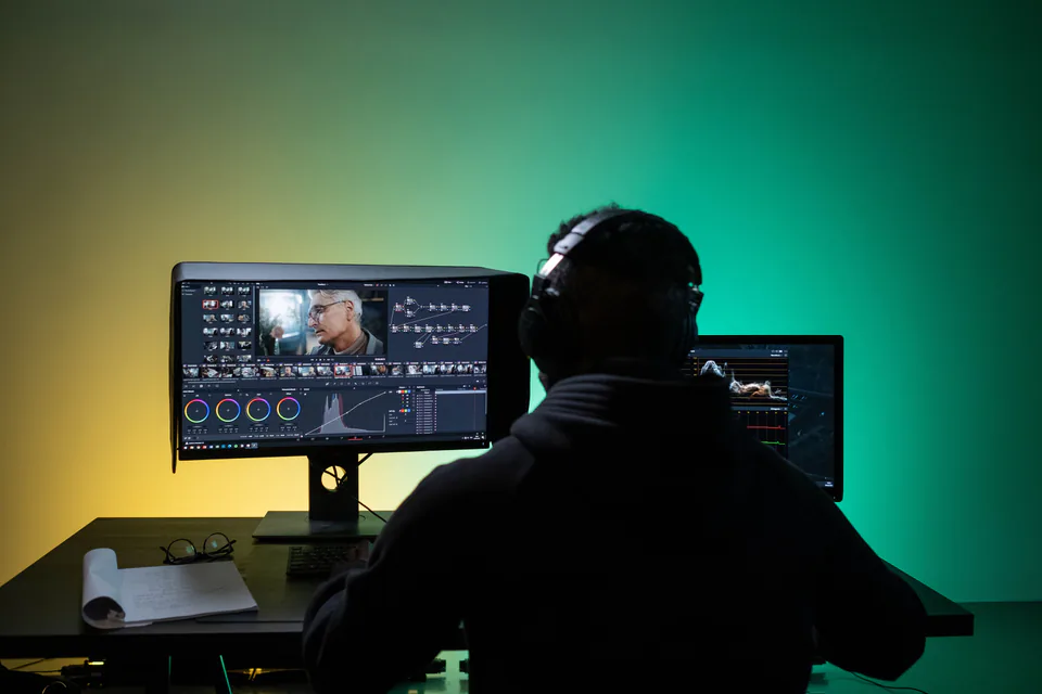 A video editor with headphones working at dual monitors against a green backlit wall