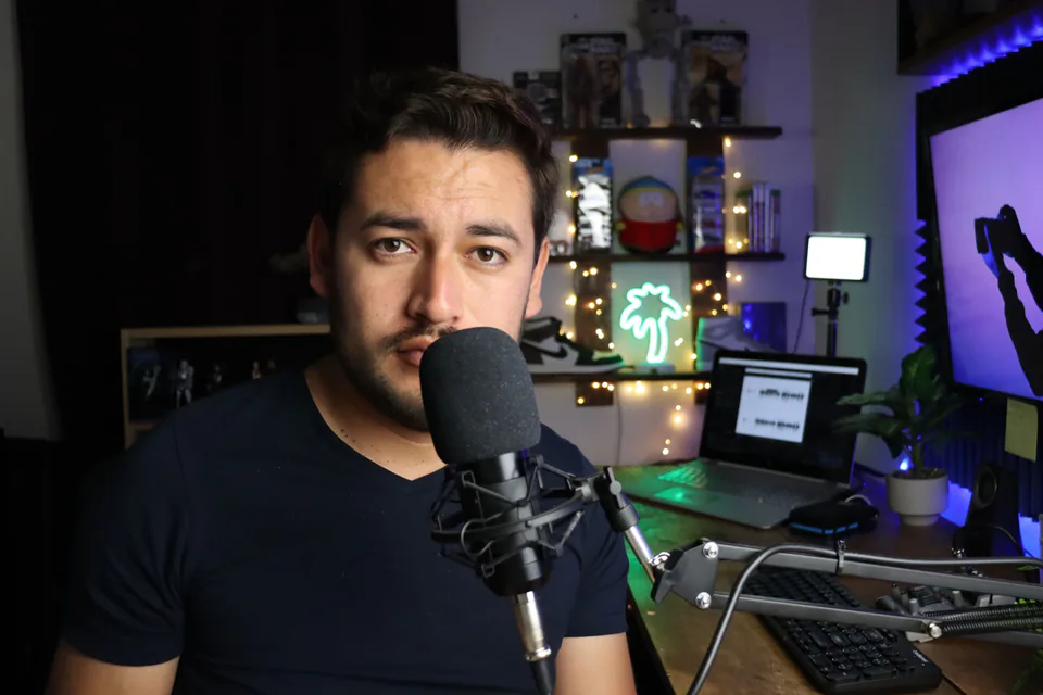 A man looking straight into the camera in front of a microphone, with a lit room behind him